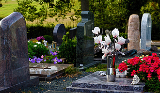 Mehrere Grabsteine auf einem Friedhof auf einer Grabplatte im Vordergrund steht eine chromfarbene hohe Vase mit Magnolienblüten darin